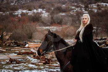 Beautiful young blonde on a crow. Woman viking with a black horse against the background of mountains