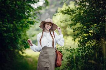 Lovely young woman with red hair, in a hat and with a backpack on the edge of the forest. Beautiful red-haired girl on a walk