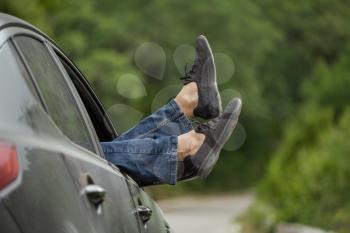 Summer road trip car vacation concept. Woman legs out the windows in car above the road in forest. Conceptual freedom, travel and holidays image with copy space.