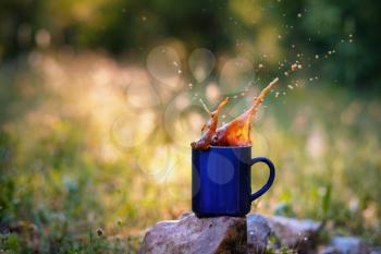 Blue mug of hot tea or coffee with milk, outdoor, the concept of travel, spray, splash. Glare on the pictures from the setting sun, pleasant bokeh