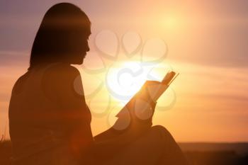 silhouette of a young beautiful woman at dawn sitting on the ground and carefully staring at the open book