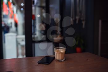 Close up of mobile phone lying on a wooden table near cup of cappuccino in coffee shop interior, leisure time concept, rest in cafe