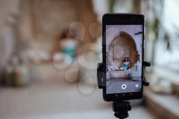 The girl is photographed on a smartphone, which stands on a tripod in Moroccan style fashion harem. Charming model posing in a red dress during her vacation to Morocco in her riad in Marrakech
