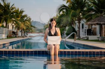 Woman using her laptop on the pool edge on a sunny day. Freelance work in tropical country