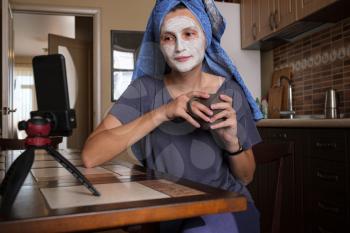 Blogger talking with followers, live streaming, looking to smartphone screen sitting on kitchen. Makes a mask of green clay. Stay at home concept for coronavirus emergency worldwide pandemic contagion