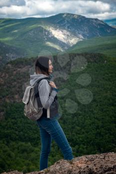 Young traveling woman with a backpack and sitting on top of the mountain cliff with relaxing mood and watching beautiful view of woods and blue sky and clouds on vacation. Asian woman travel alone.