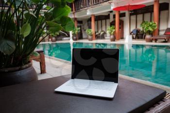 White laptop on the background of the pool in an exotic hotel. Conceptual workspace. Remote work, freelancer, internet, travel and vacation concepts.