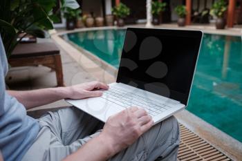 travel blogger sitting at swiming pool writing article on white laptop. Freelance remote work concept. Self employed man coding. Copy space, pool view background