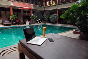 A white laptop, smartphone and mango smoothie on a sunbed against swiming pool background. A start of new day. Freelance business concept. Flexible remote working, travelling, advert and copy space