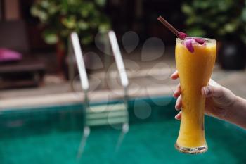 Female hand holds mango smoothie in a high glass against the background of the swimming pool