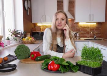 A young blonde happy lady standing in the kitchen while cooking healthy food. Healthy Lifestyle Concept. Cooking at home. broccoli, zucchini, red pepper, herbs on the table