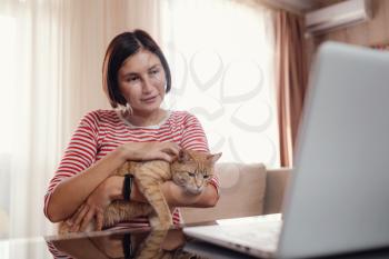 Happy young woman in a white and red tshirt works at home with a laptop and a ginger cat, remote work and education, staying home during a coronavirus