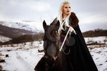 Beautiful young blonde on a crow. Woman viking with a black horse against the background of mountains. Noble Maidens
