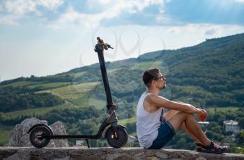 Young man riding an electric scooter on mountain range. Rest after a long trip, enjoying fresh air and beautiful scenic views. Ecological transportation concept