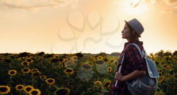 Beautiful young girl enjoying nature on the field of sunflowers at sunset. a young Asian traveller wearing a plaid shirt, hat and jeans walks in a field