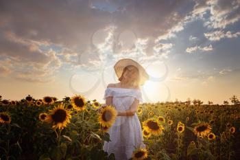 Beautiful young woman enjoying nature on the field of sunflowers at sunset. Asian girl in a cute white dress and hat enjoys summer and vacation.