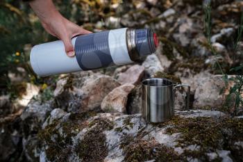 In a camping mug, a female hand pours tea from a thermos in nature. The idea and concept of a break, rest and travel