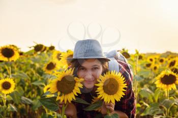 Beautiful young woman enjoying nature on the field of sunflowers at sunset. a young Asian traveller wearing a plaid shirt, hat and jeans walks in a field