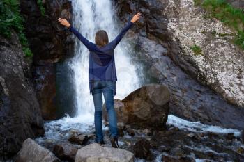 Young happy woman enjoing the waterfall. Woman standing in front of waterfall with rased hands and smile. Concept of ecotourism travel. Waterfall Shumka, Dombai, Karachay-Cherkessia, Russia