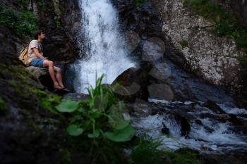 Hiker waalkinh with backpack looking at waterfall in park in beautiful nature landscape. Portrait of male adult back standing outdoor. Waterfall Shumka, Dombai, Russia