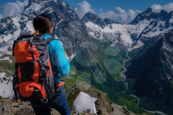 A young woman tourist in a blue blouse with an orange hiking backpack enjoys a breathtaking view of the mountains. North Caucasus, Dombai, Russia. mountaineering sport lifestyle concept