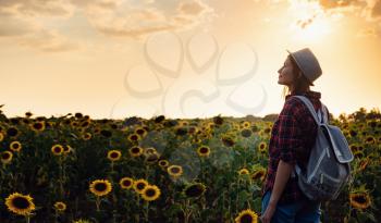 Beautiful young woman enjoying nature on the field of sunflowers at sunset. a young Asian traveller wearing a plaid shirt, hat and jeans walks in a field