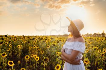 Beautiful young woman enjoying nature on the field of sunflowers at sunset. Asian woman in a cute white dress and hat enjoys summer and vacation.