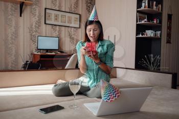 Girl celebrating birthday online in quarantine time through video call virtual party. Coronavirus outbreak 2020. Woman opening a gift and have positive emotions.