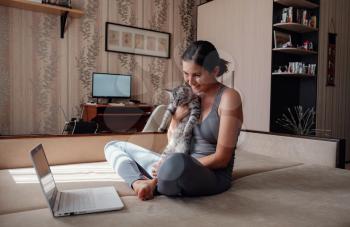 Young attractive smiling woman practicing yoga, sitting in Half Lotus exercise, wearing sportswear, meditation session, indoor full length, home interior, cat near.