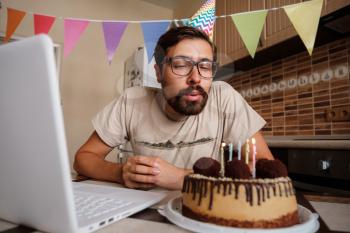 Man celebrating birthday online in quarantine time. Hipster blowing out the candle on the birthday cake and making video call.