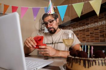 Man celebrating birthday online in quarantine time. The guy opens the box and is very happy with the gift. Communicating with friends remotely