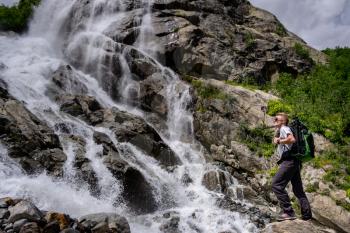 Man hiking with backpack looking at waterfall. Travel Lifestyle adventure concept, active vacations into the wild harmony with nature. North Caucasus, Dombai, Russia.