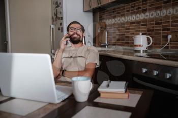 Smiling handsome freelancer working remotely from home. He is speaking on the phone. Stay at home and work from home concept during Coronavirus pandemic.