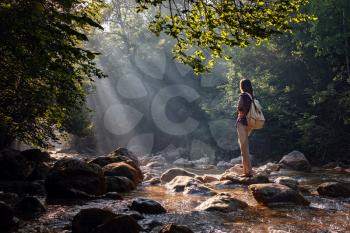 A woman explores new, magical, and fantastic places around the world, surrounded by nature and spreading her arms to breathe and relax. Female hiker crossing the forest creek.