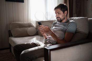 Portrait of an attractive smiling young bearded man wearing casual clothes sitting on a couch at the living room. hipster guy using social media app messenger, surfing web on phone indoors