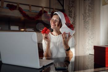 Concept of Christmas online shopping, buying gifts, congratulations over Internet. Cute girl sits with laptop, looks at the screen. Nearby gift boxes. white and red