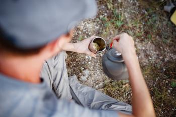 Outdoors image of young explorer man drinking hot beverage in mountains, sitting and relaxing after trekking. Traveler man in blue baseball cap holding a mug of tea after hiking. Travel people