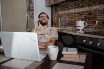 Smiling handsome freelancer working remotely from home. He is speaking on the phone. Stay at home and work from home concept during Coronavirus pandemic.