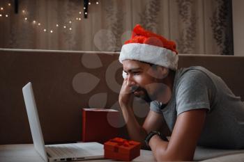 A hipster man with a red cup sitting on a sofa at home at Christmas time. Virtual Christmas house party. Online team meeting video conference calling from home.
