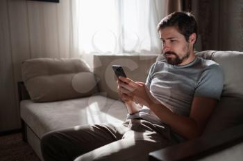 Portrait of an attractive smiling young bearded man wearing casual clothes sitting on a couch at the living room. hipster guy using social media app messenger, surfing web on phone indoors