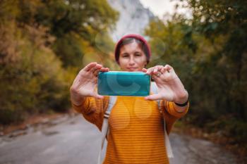 Hipster woman with backpack hold on smart phone gadget in forest. Traveler using mobile on background of mountains road. autumn lifestyle