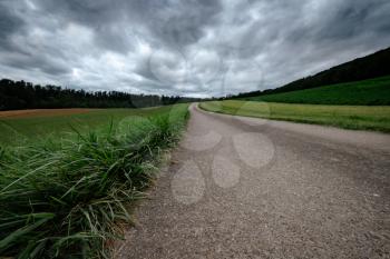 An empty country road through the green fields on a cloudly summer day. Forest in the background. Grey storm clouds.