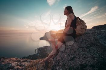 A girl sits on the edge of the cliff and looking at the sun sea and mountains. The idea and concept of recreation, freedom, summer, outdoor activities and travel