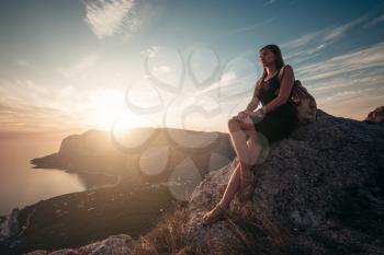 A girl sits on the edge of the cliff and looking at the sun sea and mountains. The idea and concept of recreation, freedom, summer, outdoor activities and travel