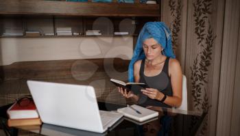 Woman with blue towel on head is working office work remotely from home. Using computer. Distance learning online education and work. Freelancer working