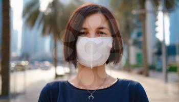 Portrait of young woman wearing in blue dress and white mask for prevent virus, walk in front of skycrapers in modern city.