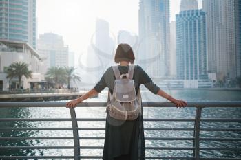 Happy young female traveler in the big city of Dubai, famous place Dubai marina. Luxury and comfortable tourism season in United Arab Emirates. Back or rear view of young woman in dress and backpack