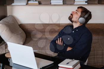 Satisfied businessman wearing headphones enjoying music with closed eyes, relaxing during break, sitting at work desk, smiling young man wearing glasses listening to favorite song