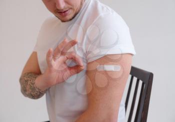 Portrait of young vaccinated man with plaster on his shoulder. Guy gesturing thumb up after coronavirus vaccination, approving of covid-19 immunization.
