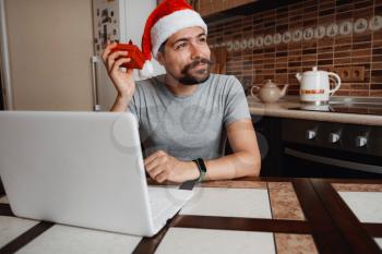A hipster man with a red cup sitting on a sofa at home at Christmas time. Virtual Christmas house party. Online team meeting video conference calling from home. Man wearing Santa hat